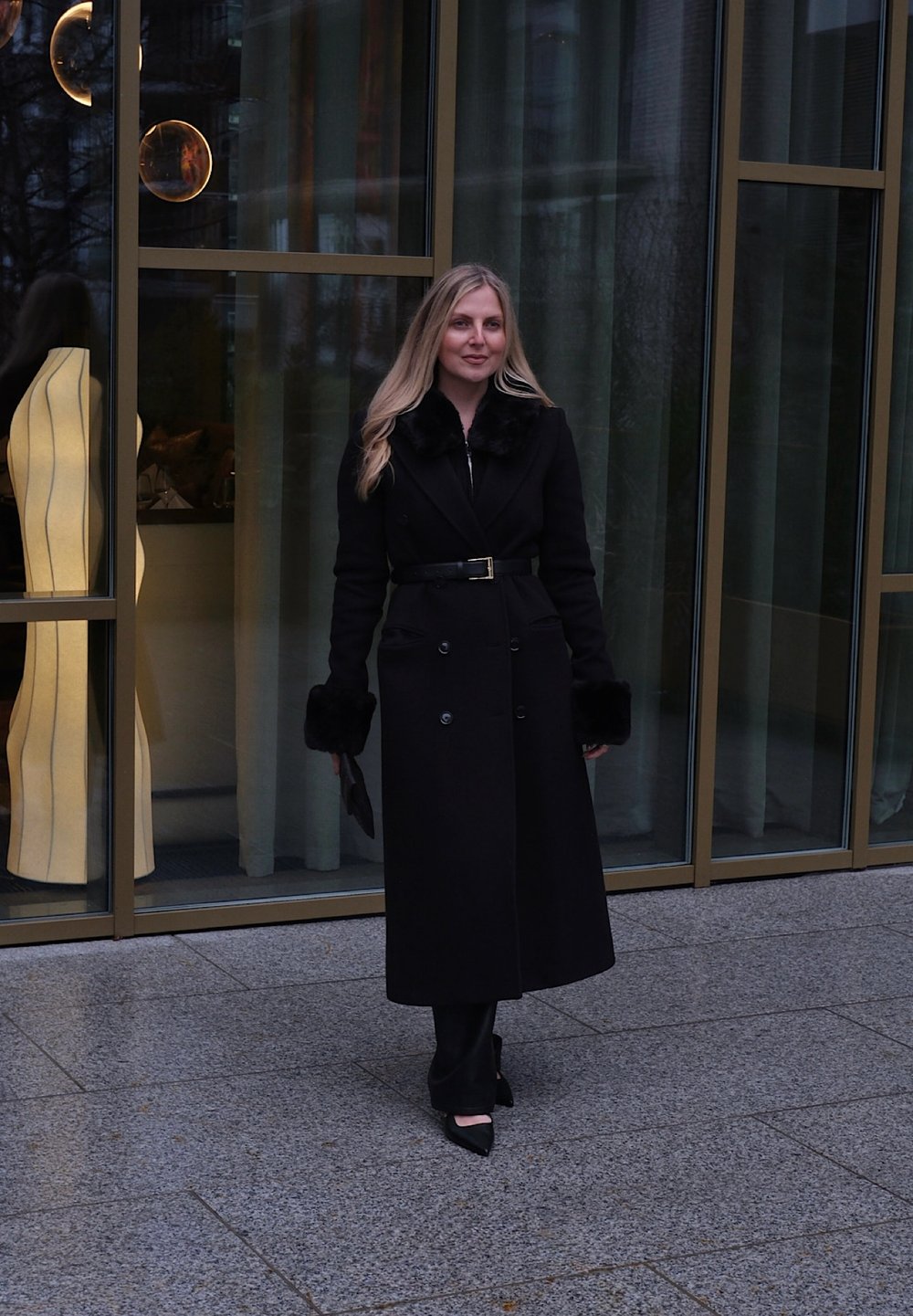 Femme aux longs cheveux blonds debout sur le trottoir portant un manteau noir ceinturé avec des manches en fourrure, un pantalon noir et des chaussures noires pointues près d'un bâtiment en verre.
