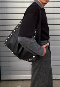 Sort lædertaske med afrundede sølvnitter langs kanterne, slasket form og struktureret, blød overflade. Fremhævet med et kontrasterende gråt stof.