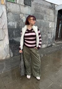 Jeune personne portant des lunettes, un pull à rayures, un pantalon olive et un gilet blanc, debout sur un trottoir mouillé à côté d'un mur en pierre, tenant un sac gris.
