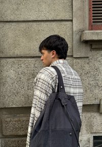 Un jeune homme aux cheveux courts et foncés, portant une chemise à carreaux et une grande sacoche noire, se tient contre un mur de pierre texturé.