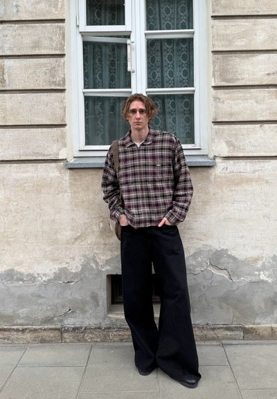 Camisa de franela de gran tamaño en tonos marrones y crema a cuadros, pantalones de pierna ancha negros, de pie contra una pared texturizada beige, con una ventana detrás.
