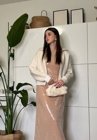 Γυναίκα που φοράει μπεζ φόρεμα με παγιέτες και λευκό αφράτο μπουφάν, κρατώντας μια εκρού τσάντα φάκελο, στέκεται κοντά σε ψηλό πράσινο φυτό και καλάθια από ψάθα.