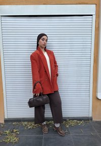 Femme portant un foulard sombre, un blazer en velours côtelé orange, un pantalon large marron, des chaussures léopard, tenant un sac à main marron, debout devant une porte métallique fermée.
