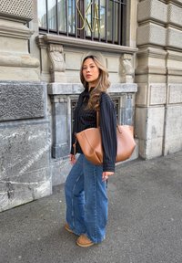 Sac à bandoulière en cuir fauve avec une forme souple et arrondie. Le modèle porte une chemise noire à fines rayures et un jean taille haute à jambes larges bleu clair dans la rue.