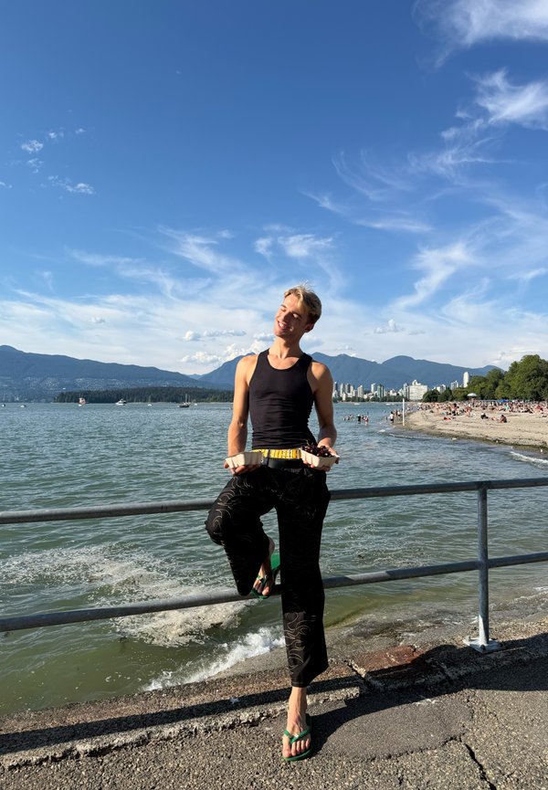 Person in schwarzem Tanktop und Hose, die Essensbehälter hält, steht auf einem Bein am Geländer mit Ozean, Strand und Bergen im Hintergrund.