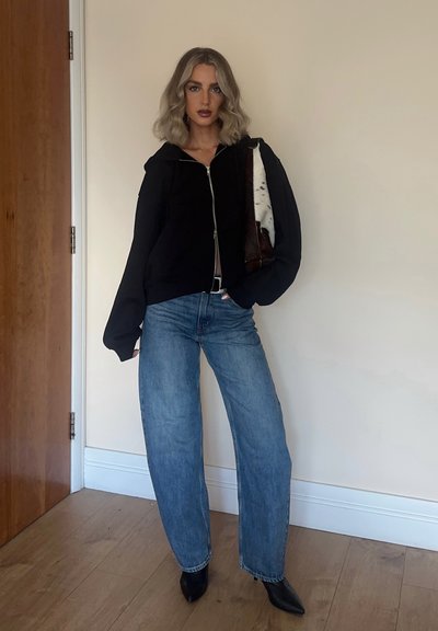 Sudadera cropped negra con cremallera y mangas oversized, combinada con jeans de pierna ancha azul claro y botas negras de tacón puntiagudo. Bolso de diseño marrón y blanco.