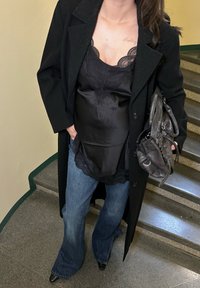 Camisole noire à couches avec bordure en dentelle, manteau noir oversize, jeans évasés bleus et talons noirs pointus, tenant un sac noir texturé.