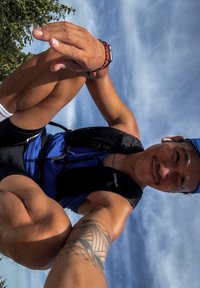 Idrettslig mann iført en blå singlet og svarte shorts, med en svart ryggsekk. Merkbar tatovering på armen hans. Himmel synlig i bakgrunnen.