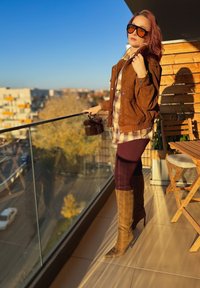 Giacca in camoscio marrone sopra una camicia a quadri, leggings bordeaux e stivali marroni con tacco fino al ginocchio. Accessori includono occhiali da sole neri oversize e una borsa di pelliccia.