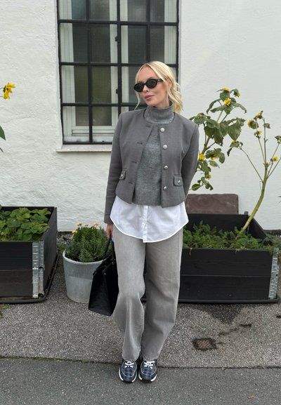 Chaqueta gris corta con botones de metal, cuello alto gris, camisa blanca, pantalones gris claro, bolso negro y zapatillas con estampado. Entorno al aire libre.