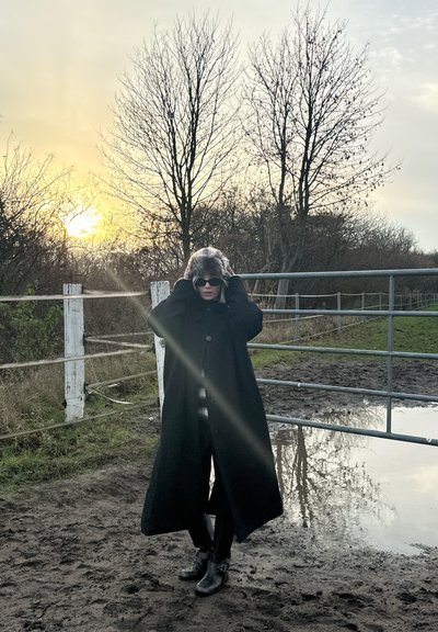 Abrigo negro largo, sombrero gris peludo, gafas de sol oscuras, de pie en un área fangosa cerca de una puerta. El fondo presenta árboles sin hojas y una puesta de sol.