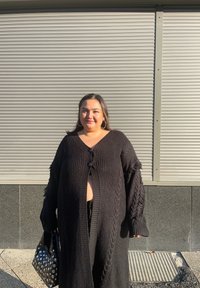 Černý pletený cardigan s výstřihem do V, nadměrným střihem a texturovaným vzorem copánků. Má zapínání na knoflíky vpředu a třásně na rukávech. Černá kabelka se stříbrnými doplňky.