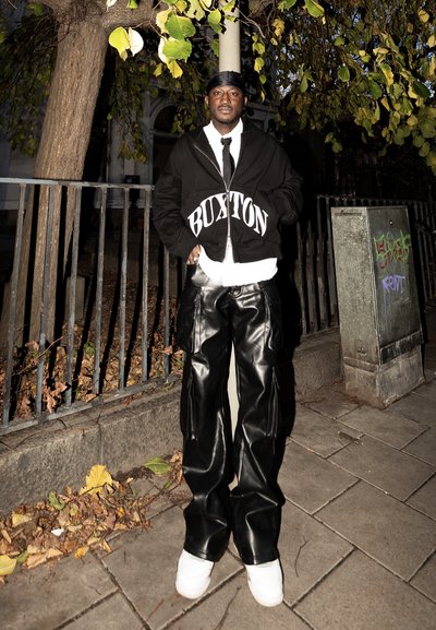 Hombre de pie en la acera por la noche con durag negro, sudadera con capucha negra "Buxton", pantalones de cuero negro, camisa blanca, corbata y zapatillas blancas.