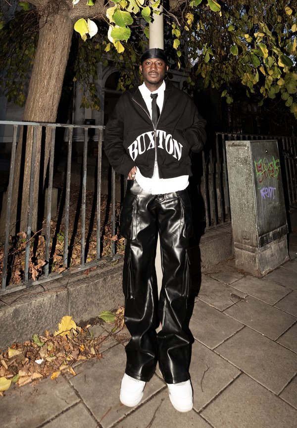 Un homme se tient sur le trottoir la nuit, portant un durag noir, un sweat à capuche noir "Buxton", un pantalon en cuir noir, une chemise blanche, une cravate et des baskets blanches.