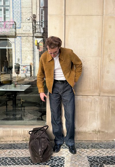 Hombre con chaqueta marrón y pantalones oscuros se apoya contra una pared beige, mirando hacia abajo una bolsa de lona marrón sobre una acera con patrón cerca de una ventana de vidrio.