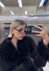 Sort faux fur frakke med bred krave, tilbehør der inkluderer tynde, rektangulære bordeaux briller og sølvringe, som reflekterer en metallisk baggrund.