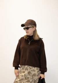 Femme portant une casquette marron, des lunettes de soleil et un pull à fermeture éclair associé à une jupe beige camouflage, tenant un sac noir sur un fond uni.
