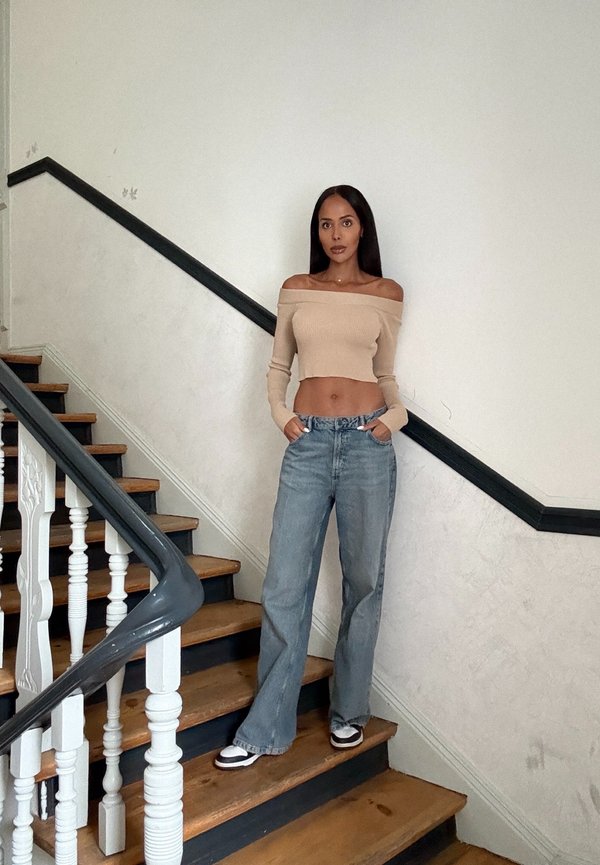 Top corto de canalé beige con los hombros al descubierto, combinado con jeans de tiro alto de color azul claro. La modelo está de pie en unas escaleras de madera con barandilla blanca.