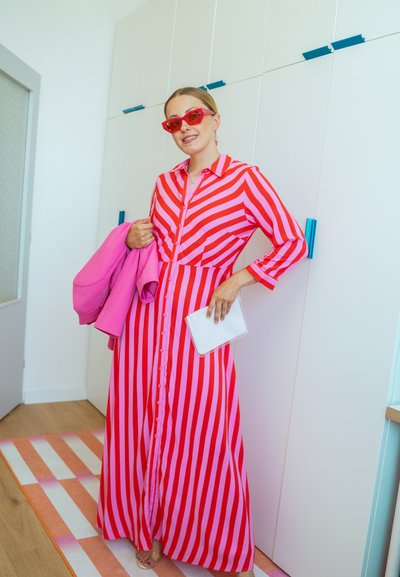 Vestido maxi a rayas rojas y rosas con mangas largas y botones en la parte delantera, combinado con una chaqueta rosa, un clutch blanco y gafas de sol rojas.