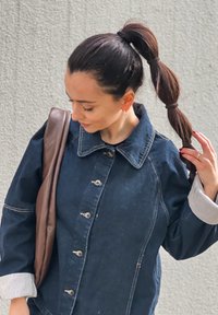 Mornarsko modra jeans jakna s ovratnikom, gumbastim zapiranjem in črtastimi manšetami; elegantna rjava torba za rame; lasje zvezani v visokem repu.