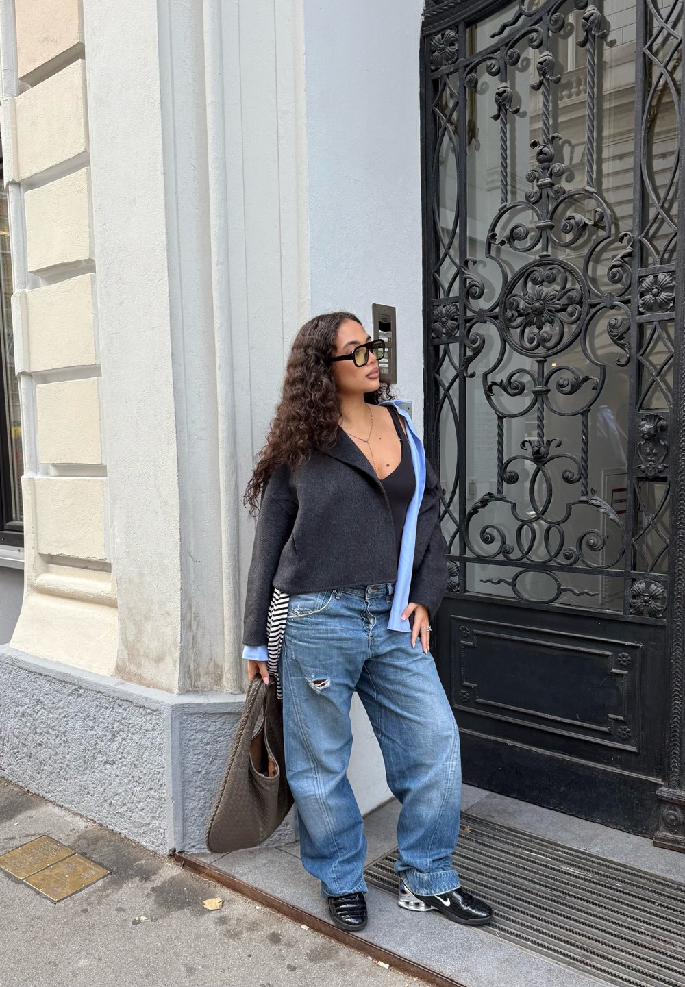 Sweater bicolor em preto e azul, jeans azuis esfarrapados, óculos oversized pretos e bolsa texturizada castanha; de pé ao lado de uma porta de metal ornamentada.