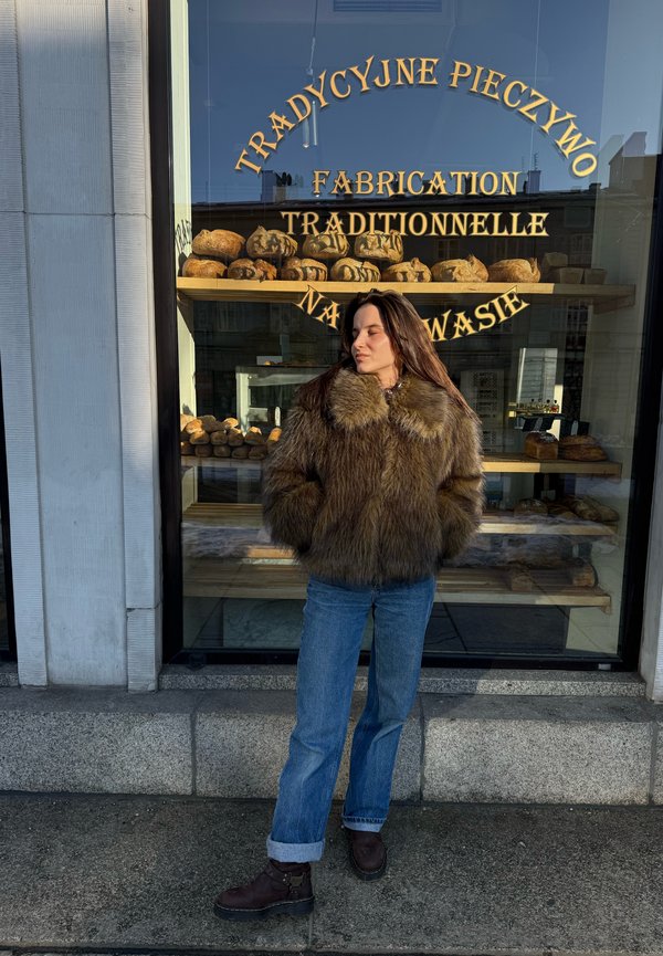 Kvinde iført en brun pelsjakke og blå jeans står med hænderne i lommerne foran et bagerivindue, hvor der vises brødflutes.