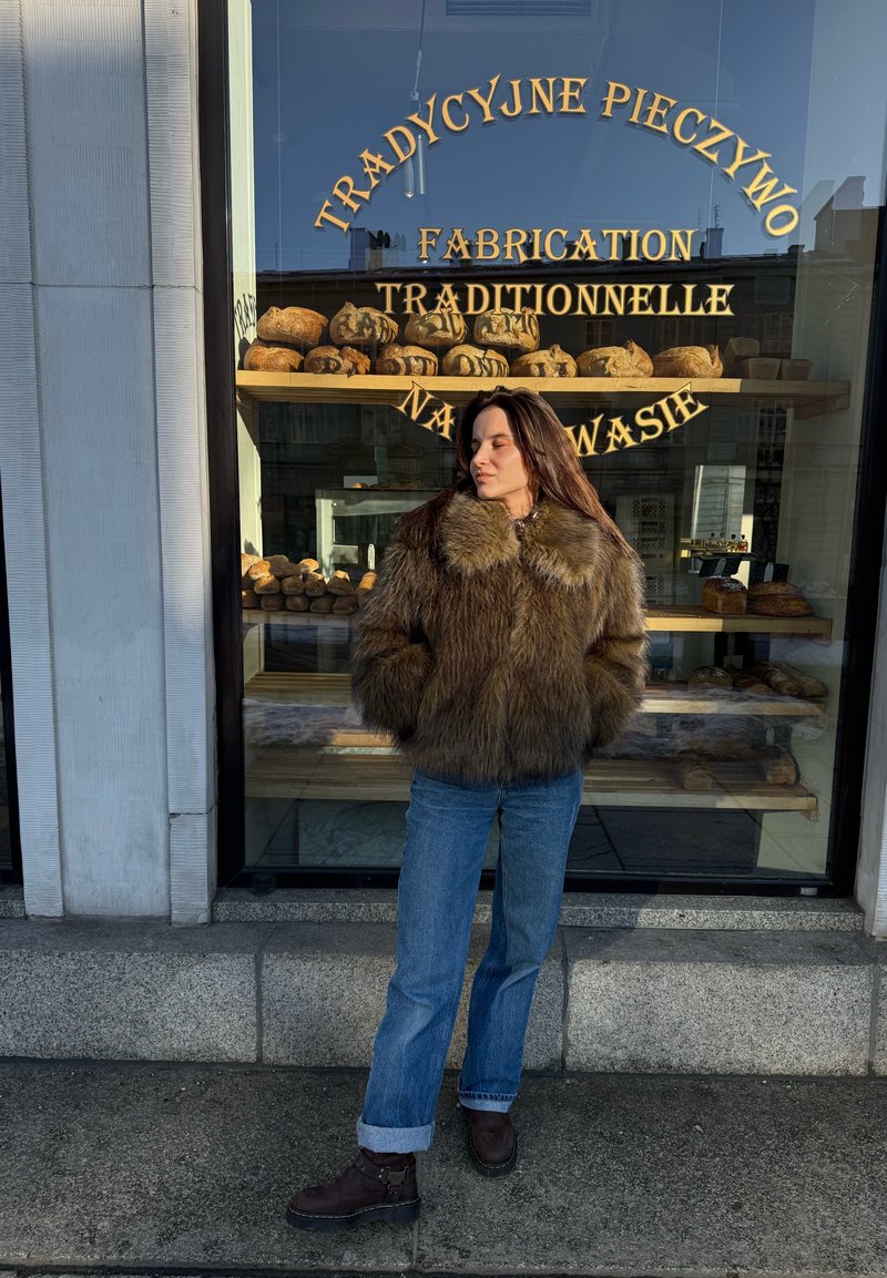 Kvinde iført en brun pelsjakke og blå jeans står med hænderne i lommerne foran et bagerivindue, hvor der vises brødflutes.
