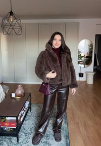 Femme portant une veste en fausse fourrure marron, un pantalon en cuir et des mocassins, tenant un sac à main violet, debout dans un salon moderne.