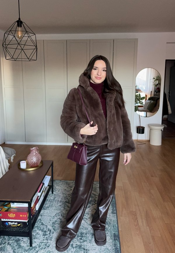 Femme portant une veste en fausse fourrure marron, un pantalon en cuir et des mocassins, tenant un sac à main violet, debout dans un salon moderne.