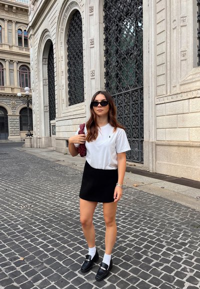 Camiseta blanca, falda corta negra, mocasines negros y calcetines blancos. Lleva una bolsa burdeos. Calle adoquinada y edificio ornamentado de fondo.