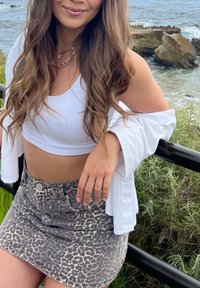 White ribbed crop top, light long-sleeve shirt draped, and gray-leopard print denim skirt. Close-up on the torso, hand resting on a railing.