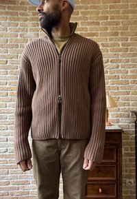 Suéter marrón de canalé con cremallera y cuello alto, combinado con pantalones marrones. Tejido texturizado y ajuste ceñido.