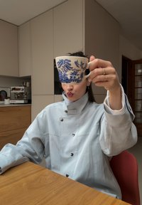 Tasse en céramique avec un motif floral bleu tenue devant une personne. La personne porte un manteau gris clair boutonné. Surface de table en bois visible.