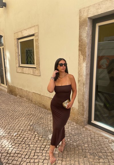 Vestido midi ajustado marrón con una textura suave. Accesorios incluyen un bolso dorado de mano y gafas de sol. De pie en una calle de adoquines.