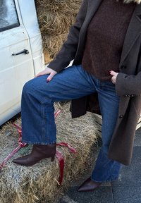 Personne portant un manteau marron, un pull bordeaux, un jean large bleu et des bottines marron à talons se tient avec le pied posé sur une botte de foin attachée par un ruban rouge.