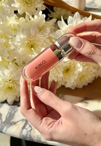 Προϊόν χειλιών KIKO Milano σε διαφανή κυλινδρική συσκευασία με ασημένιες λεπτομέρειες, που περιέχει μια κρεμώδη φόρμουλα σε ανοιχτό ροζ χρώμα, κρατημένη μπροστά από λευκά λουλούδια.