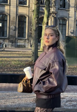 Mujer con chaqueta de cuero marrón sosteniendo una taza de café y un bolso de diseño, de pie al aire libre cerca de árboles y un edificio histórico a la luz del día.