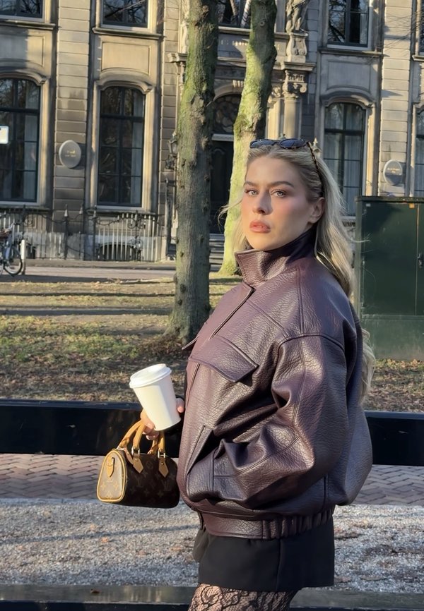 Frau in brauner Lederjacke, die eine Kaffeetasse und eine Designertasche hält, steht tagsüber draußen in der Nähe von Bäumen und einem historischen Gebäude.