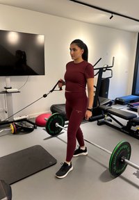Femme en tenue de sport bordeaux tenant une poignée de câble de résistance, debout dans une salle de sport à domicile avec des poids, du matériel d'exercice et un écran de télévision.