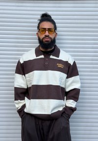 Ruskean ja vaalean raidallinen neulottu poolopaita, jossa on kaulus, kolme nappia ja kullalla kirjailtu teksti "laidback stories". Yhdistetty tummiin housuihin.