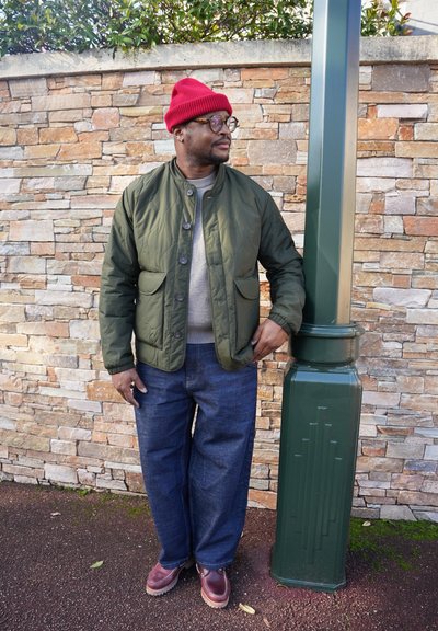 Chaqueta acolchada verde con dos bolsillos, suéter gris, jeans anchos azules y gorro de punto rojo. De pie junto a un farol verde.