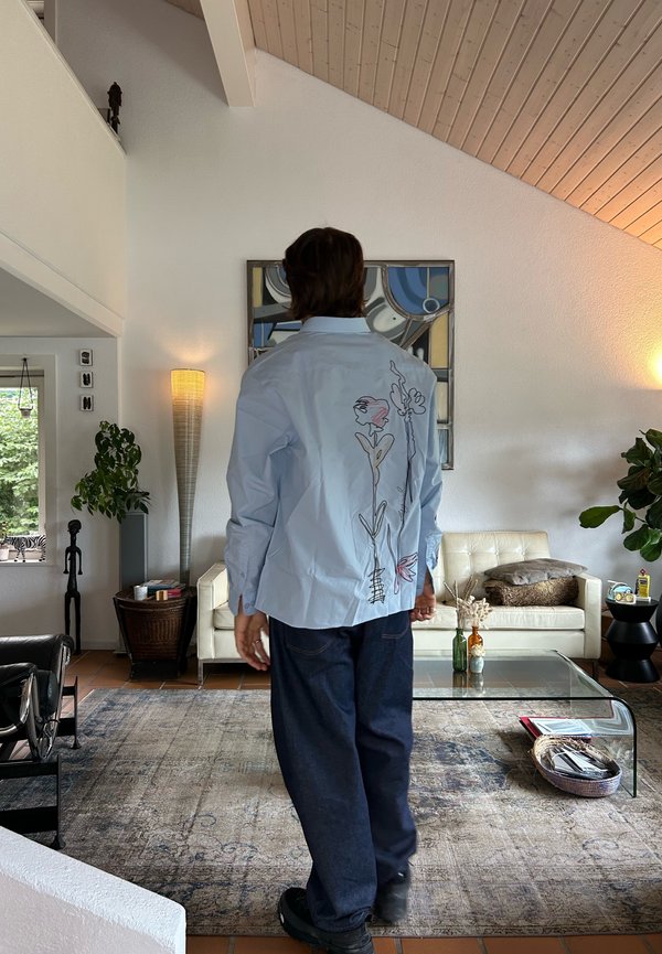 Persona che indossa una camicia blu chiaro con arte floreale a linea sul retro, pantaloni scuri, in piedi in un soggiorno moderno con piante e un tavolino da caffè in vetro.