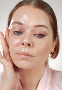 Zbliżenie na twarz z lśniącą, nawilżoną skórą. Jasnobrązowe włosy, przenikliwe niebieskie oczy i kolczyk w przegrodzie nosa. Miękki różowy satynowy top.