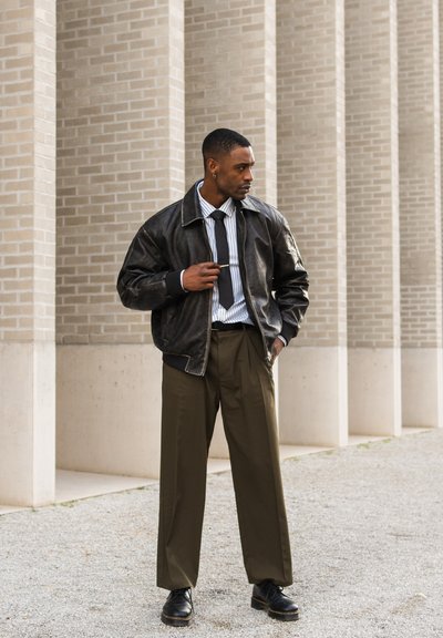 Chaqueta bomber de piel negra, camisa a rayas blanca y azul, corbata negra, pantalones de pierna ancha verde oliva, zapatos negros, pose de pie. Fondo texturizado.