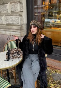 Cappello e borsa con stampa leopardata in materiale morbido; lungo cappotto nero; dolcevita nero aderente; pantaloni grigi a gamba larga; tavolo rotondo con bordo piastrellato.