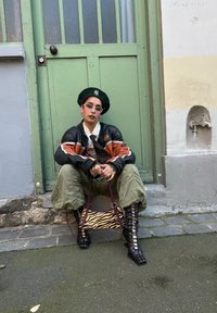 Henkilö mustassa ja oranssissa nahkatakissa, oliivinvihreissä cargo-housuissa, mustissa nauhaketjuilla varustetuissa korkokengissä, vihreässä barettissa, pitämässä tiikeriraidoitettua laukkua.