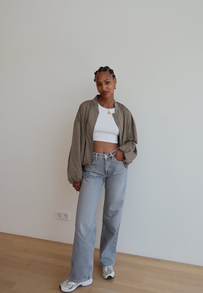 Chaqueta bomber caqui, top corto de canalé blanco, vaqueros de tiro alto azul claro y zapatillas gris-blancas sobre un fondo liso.