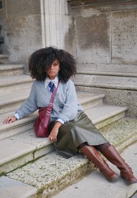 Cardigan in maglia grigia sopra una camicia con colletto, gonna plissettata verde, borsa a tracolla bordeaux e stivali marroni fino al ginocchio, seduta su gradini di pietra.