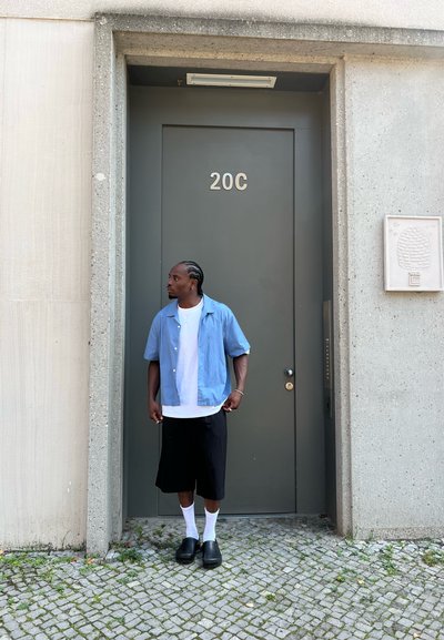 Outfit informal que presenta una camisa de manga corta azul claro, una camiseta blanca, pantalones cortos negros hasta la rodilla, calcetas blancas y zuecos negros. De pie junto a una puerta gris.