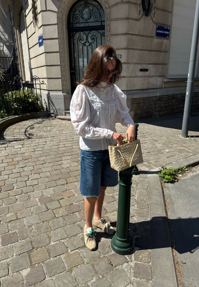 Bolso de mano beige de tejido con asas cortas, combinado con una blusa blanca con bordado y pantalones cortos de denim, sobre un pavimento de adoquines.